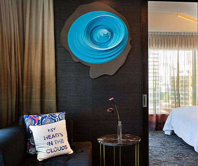 A large circular emblem mounted on a wall above a marble coffee table and cushioned bench. An open doorway leads to a bedroom